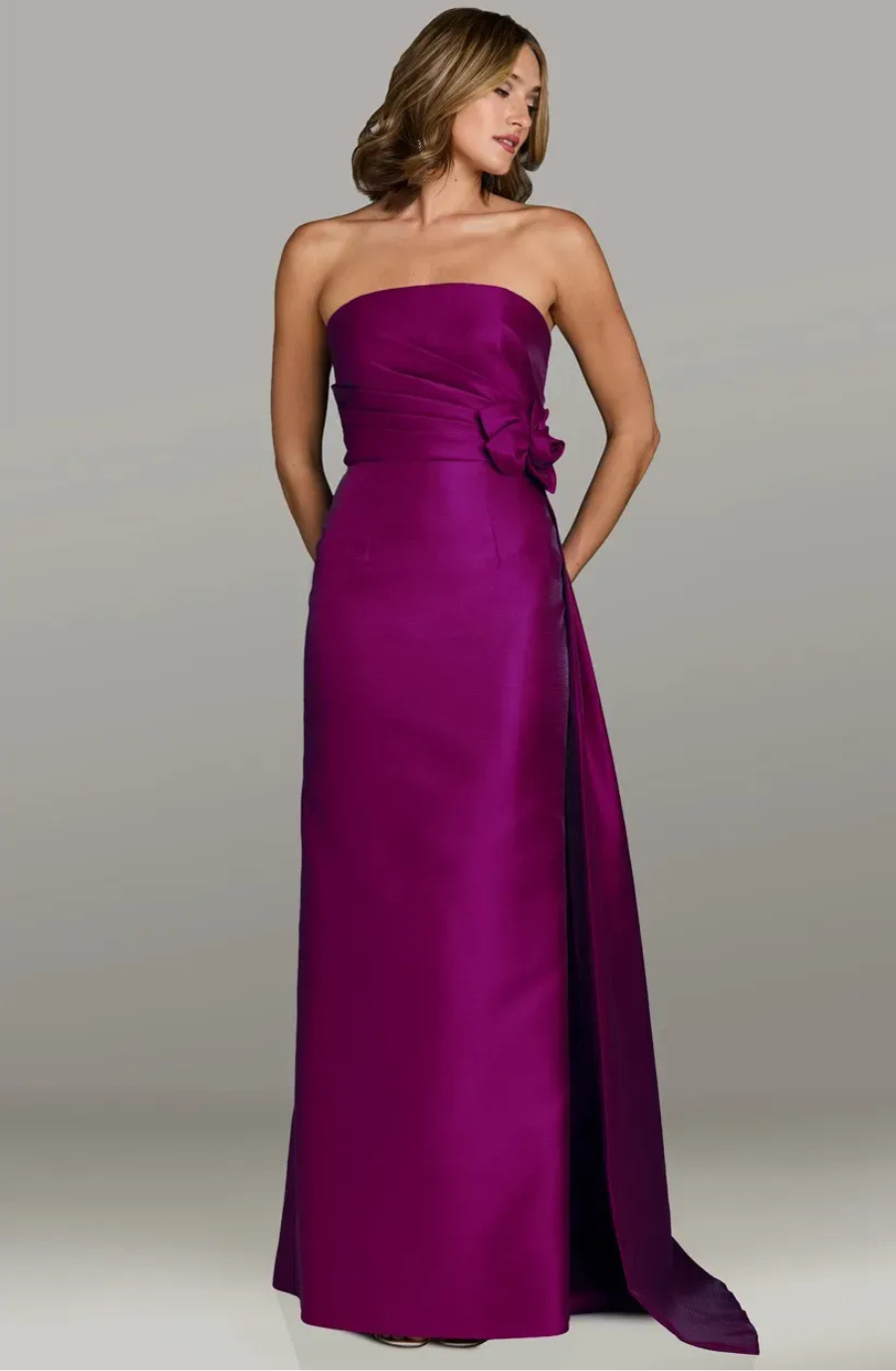 A woman in a strapless, floor-length magenta gown with a floral accent stands gracefully against a gray backdrop, exuding elegance and poise.