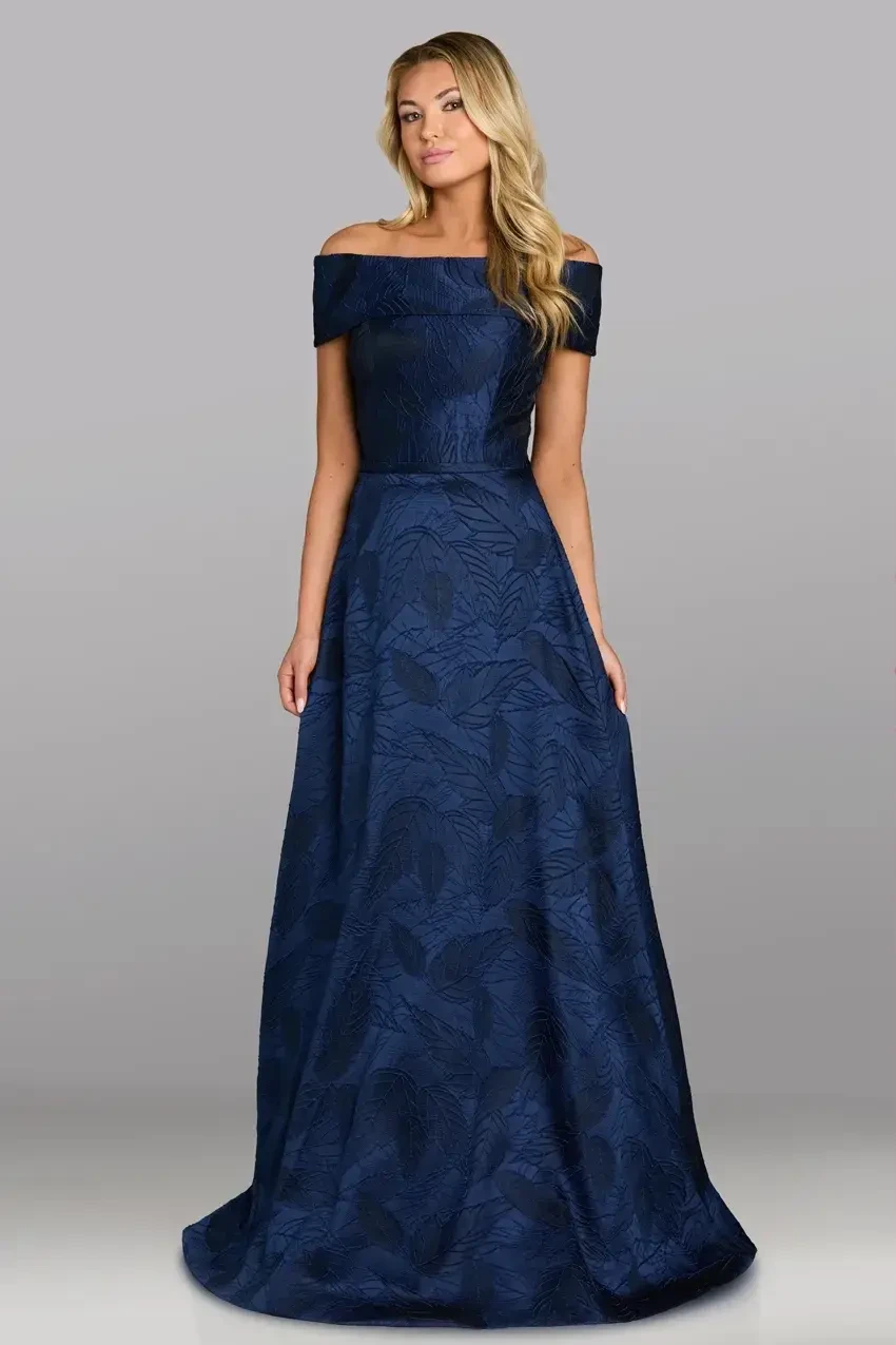Woman wearing a floor-length, off-the-shoulder navy blue gown with a subtle leaf pattern, standing against a gray background. She looks elegant and poised.