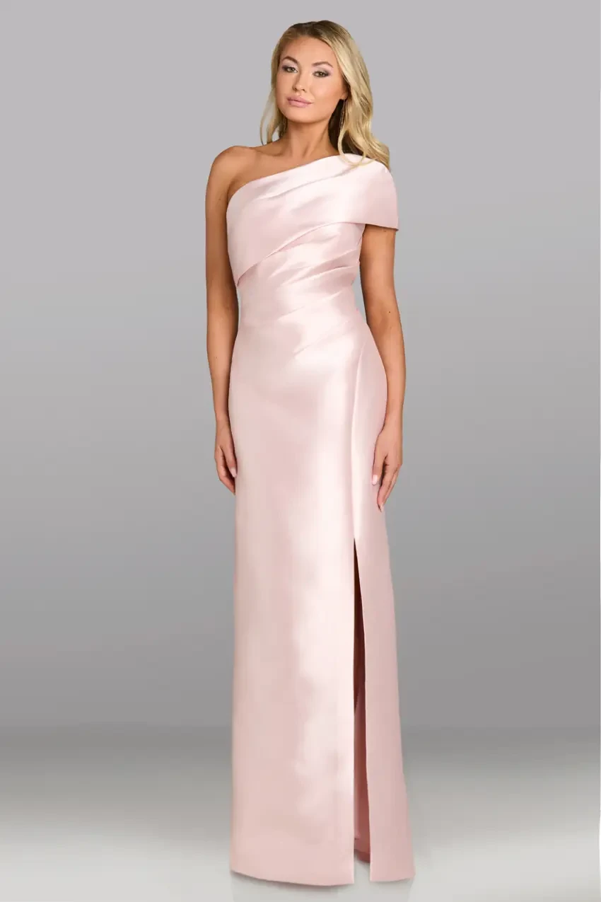 A woman stands in a sleek, pale pink, one-shoulder gown with a thigh-high slit, exuding elegance. The backdrop is a simple gray, highlighting the dress.