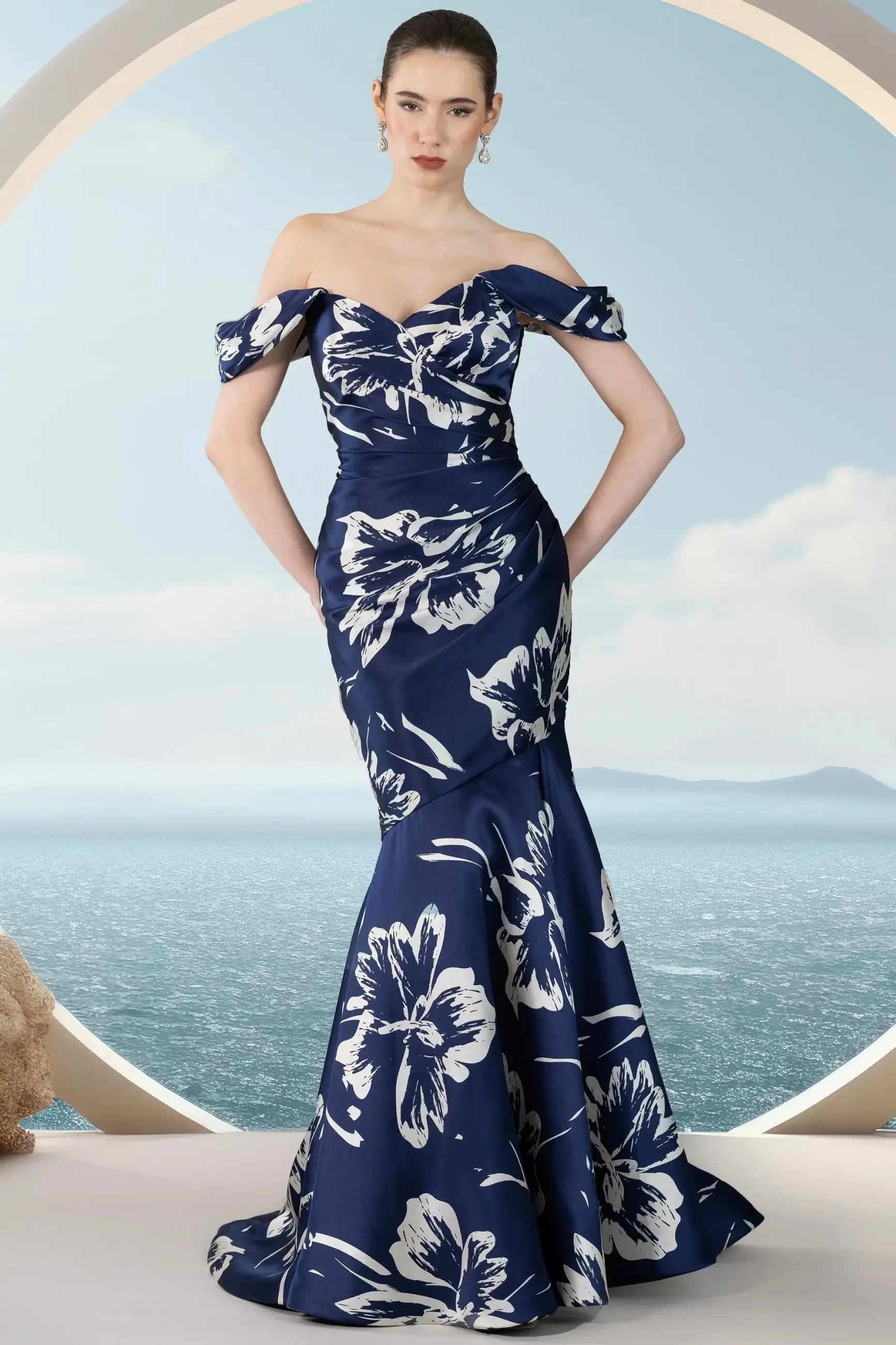 A model wearing an off-the-shoulder floral gown, standing in front of a scenic backdrop with the ocean and sky.