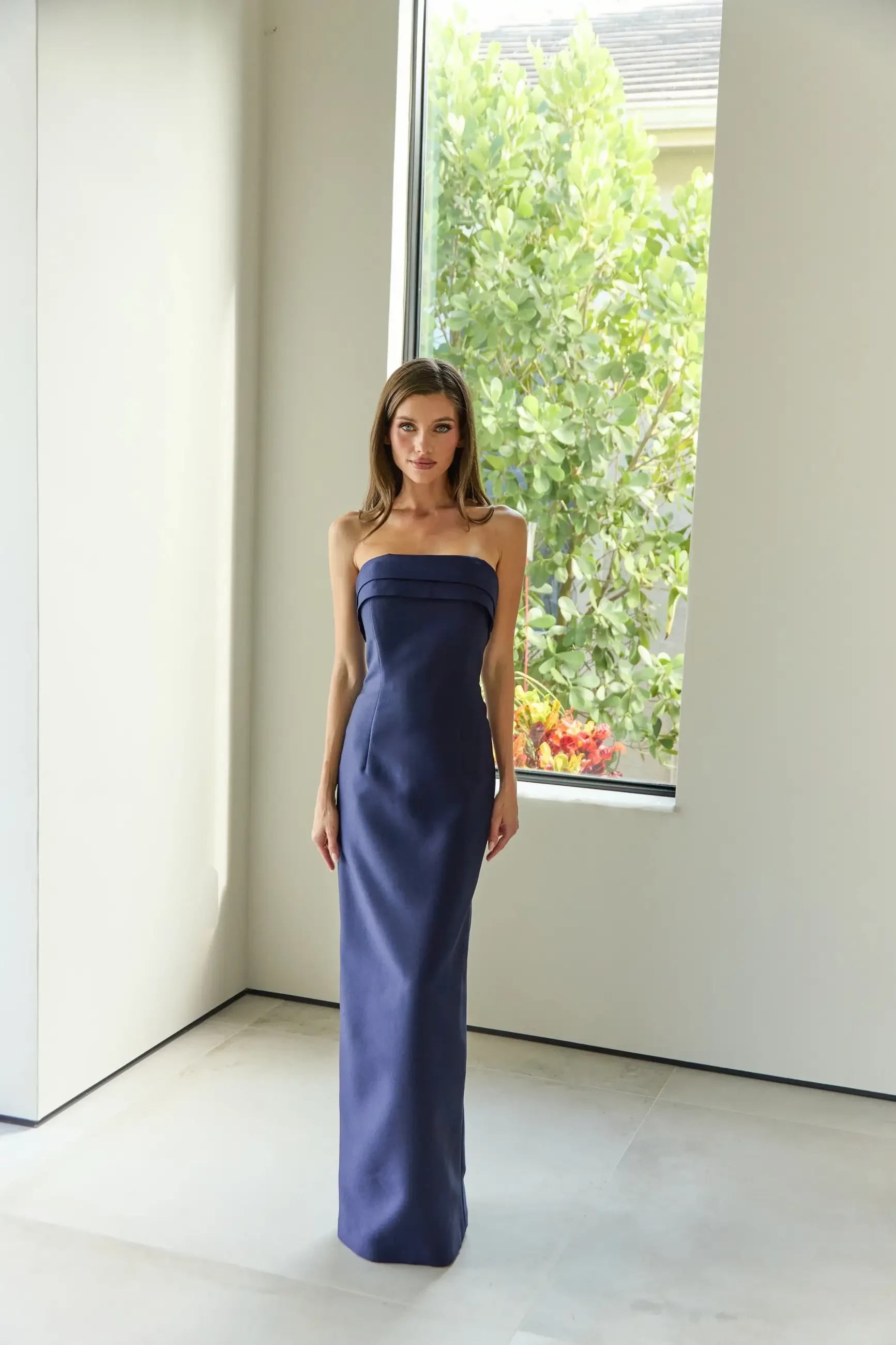 A woman wearing a strapless blue evening gown stands in front of a large window with greenery visible outside.