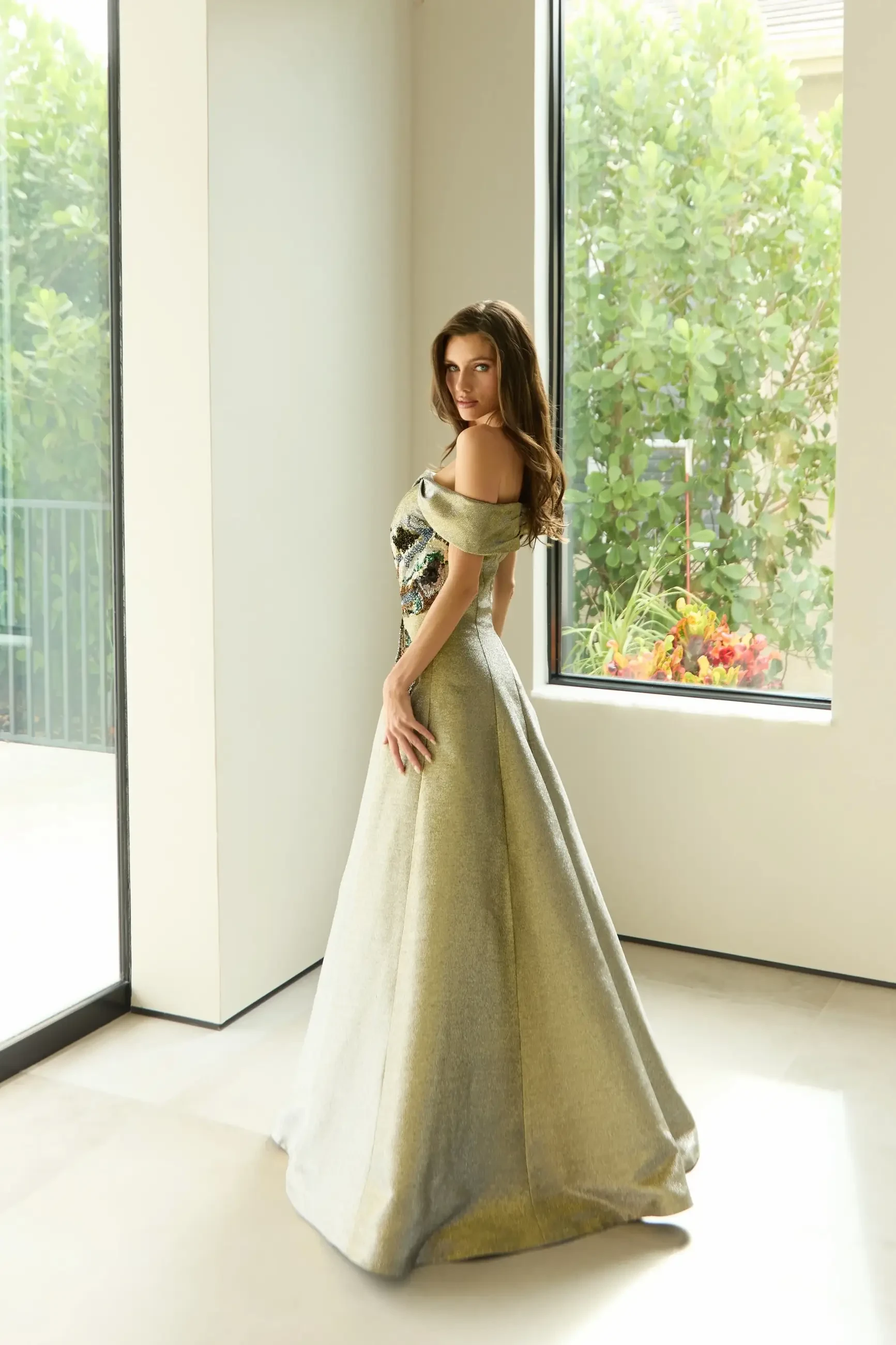 A woman in an elegant, off-shoulder gown stands gracefully with her back turned, looking over her shoulder in a well-lit room with large windows and greenery outside.