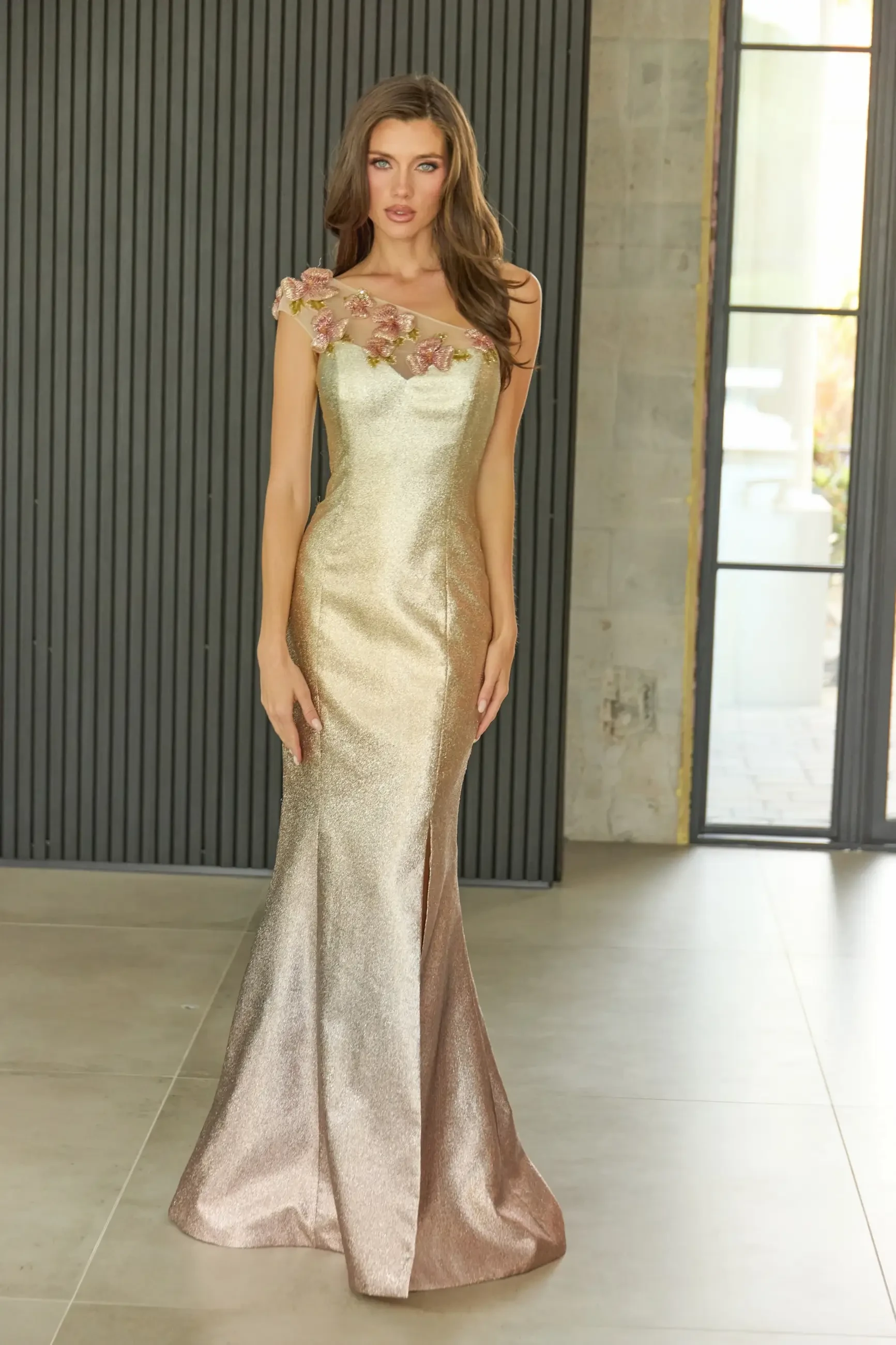A model wearing a fitted, elegant gown with floral embellishments and a gradient color scheme, standing in a modern interior space.