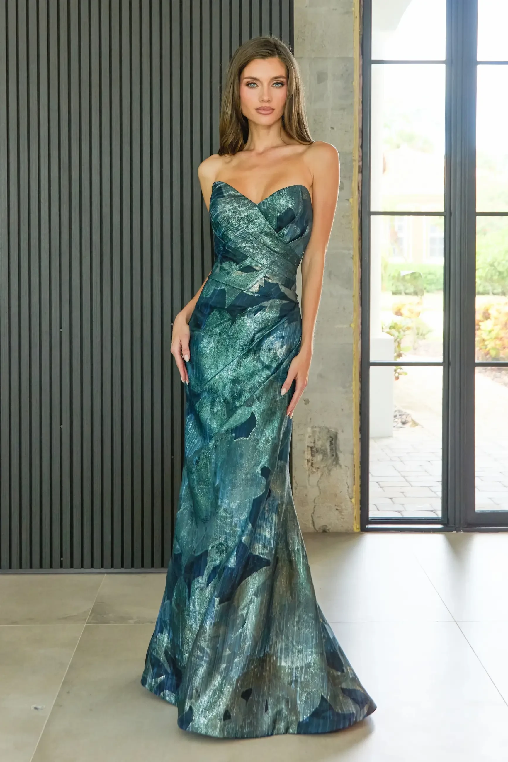 A model wearing a strapless, floor-length gown with a blue and green abstract pattern, standing against a backdrop of large windows and vertical slatted walls.