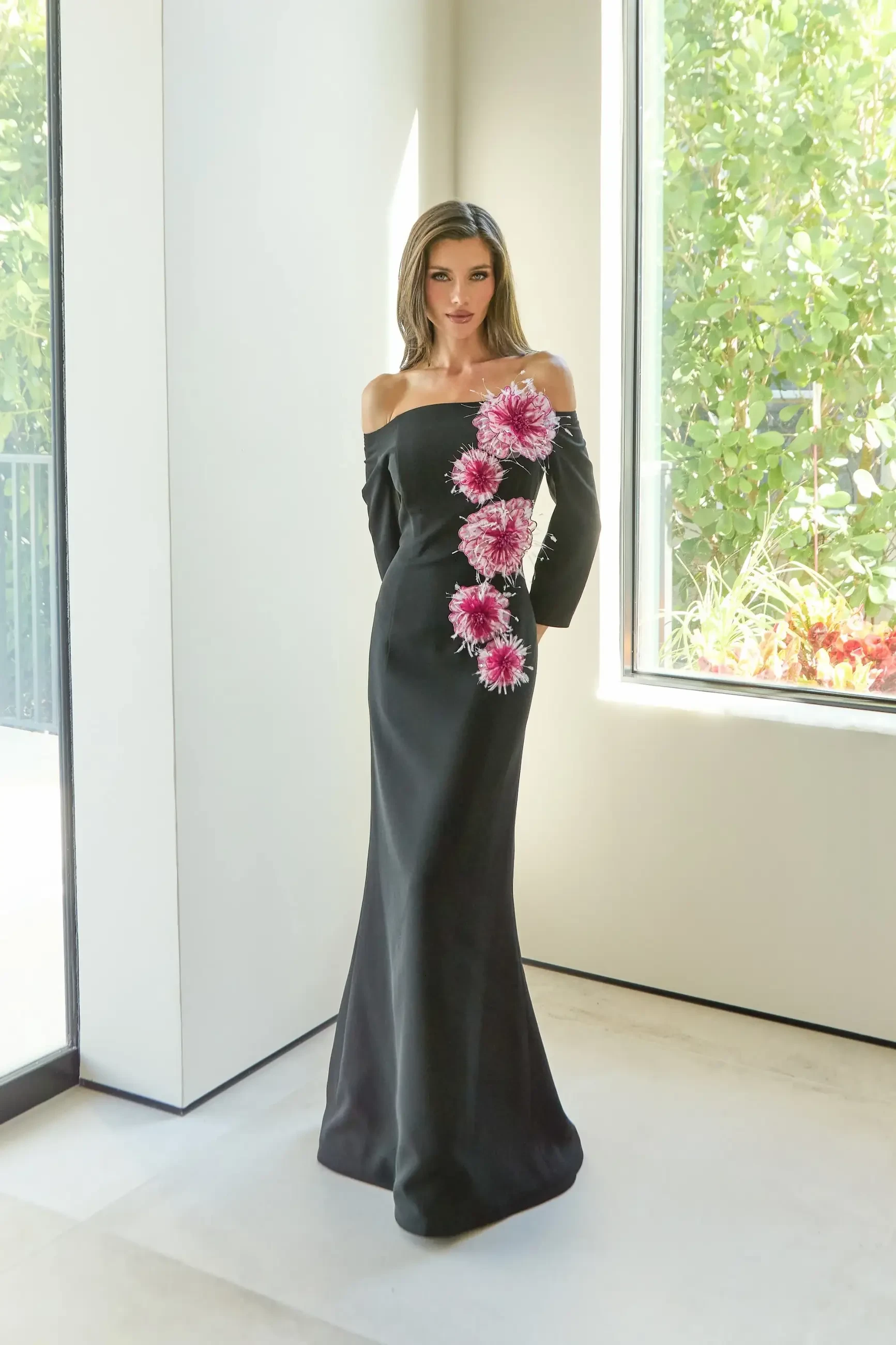 A woman wearing an elegant, off-the-shoulder black dress adorned with pink flowers on one side, standing in a well-lit interior near a window.
