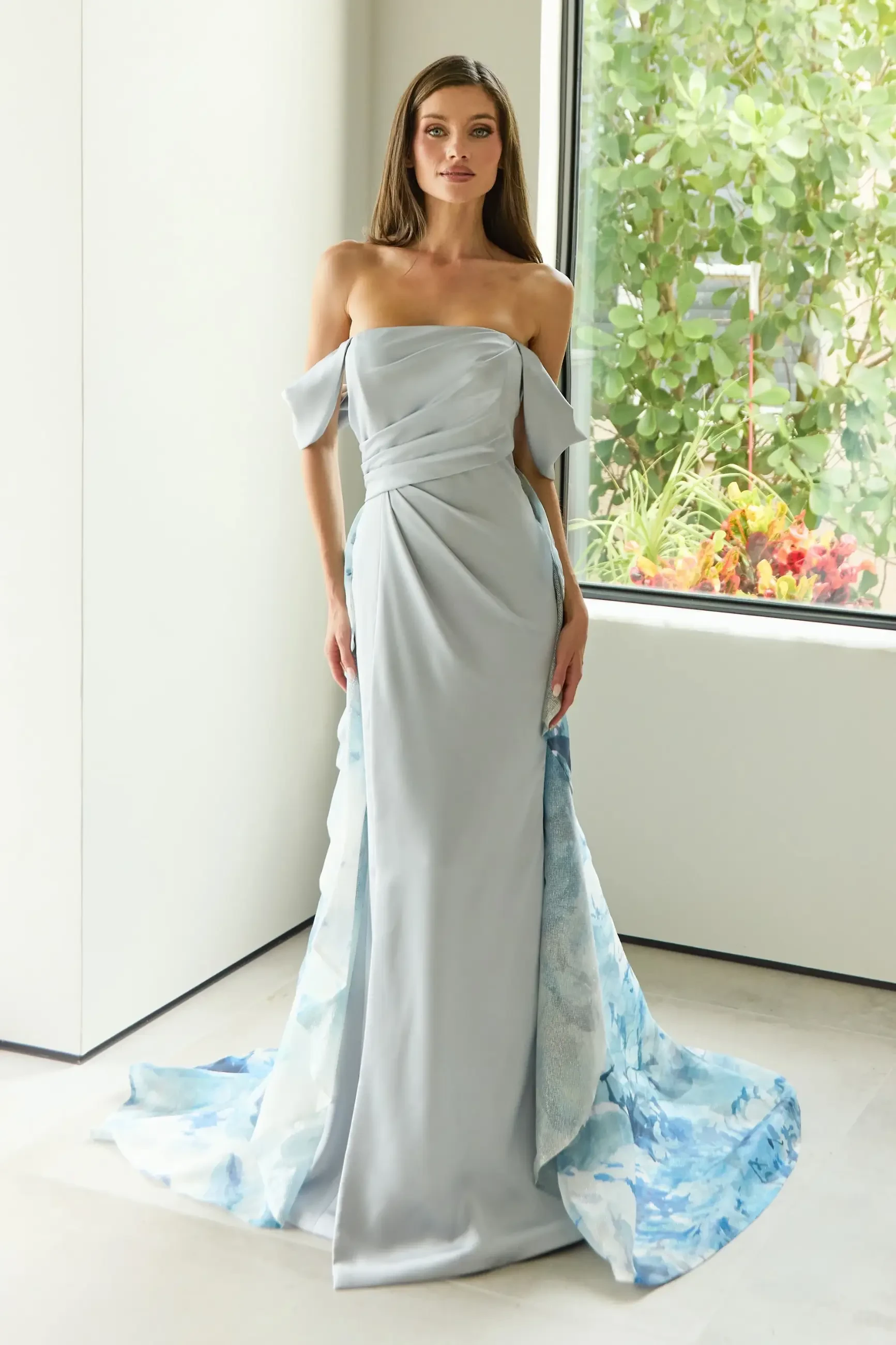 A woman wearing a strapless, off-the-shoulder gray gown with a flowing blue floral train, standing in an indoor setting with greenery and colorful flowers visible through a large window.