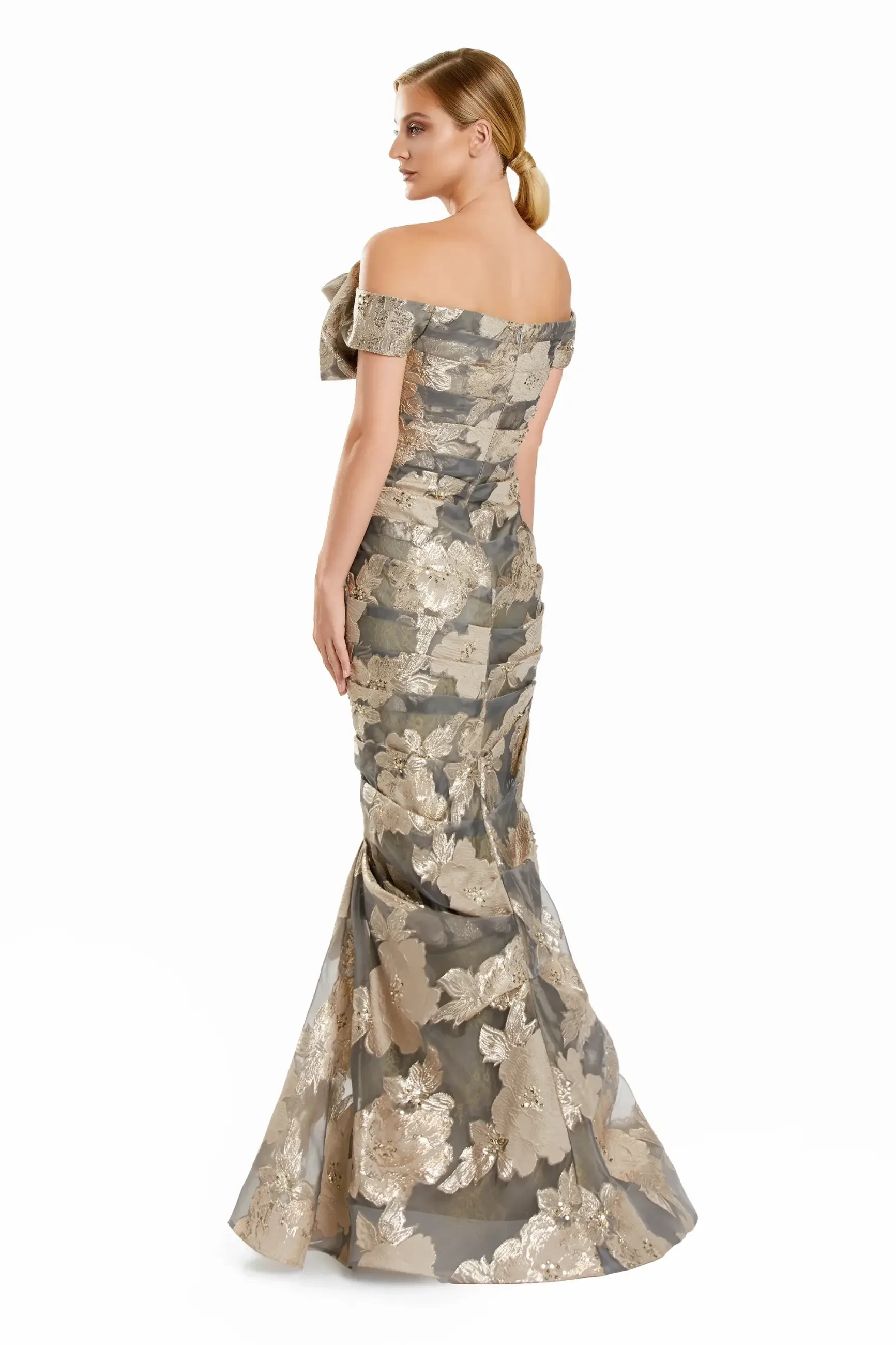 A woman in an elegant, off-shoulder gown with gold floral patterns stands against a white background, exuding a sophisticated and graceful tone.