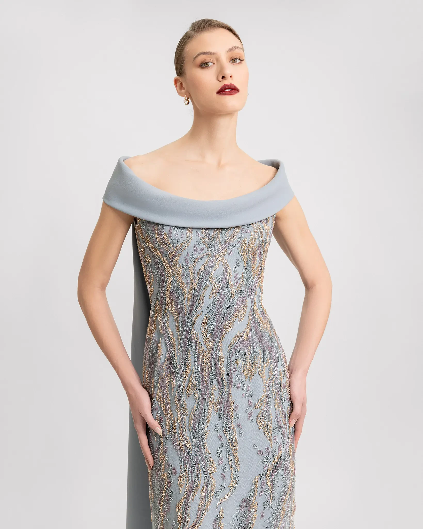 Model is wearing a sleek grey embroidered off the shoulder evening gown