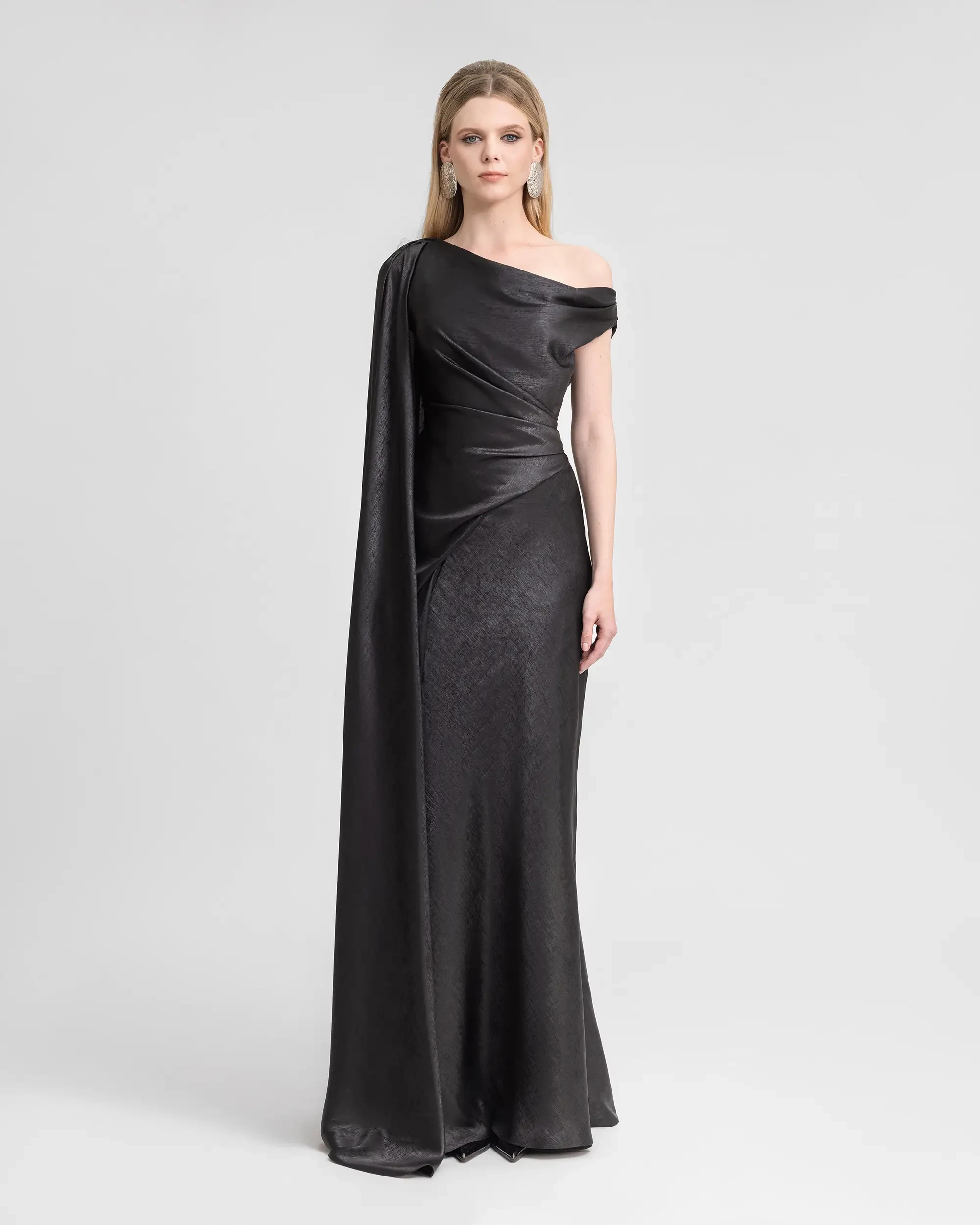Model is wearing a black asymmetrical neckline evening gown with satin bodice and textured skirt and a long cape on one arm