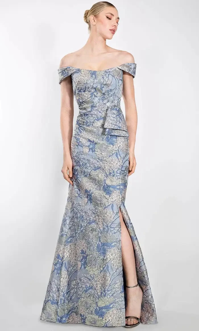 A woman in an elegant off-the-shoulder gown with a blue and silver floral pattern, featuring a thigh-high slit. She stands gracefully against a neutral background.
