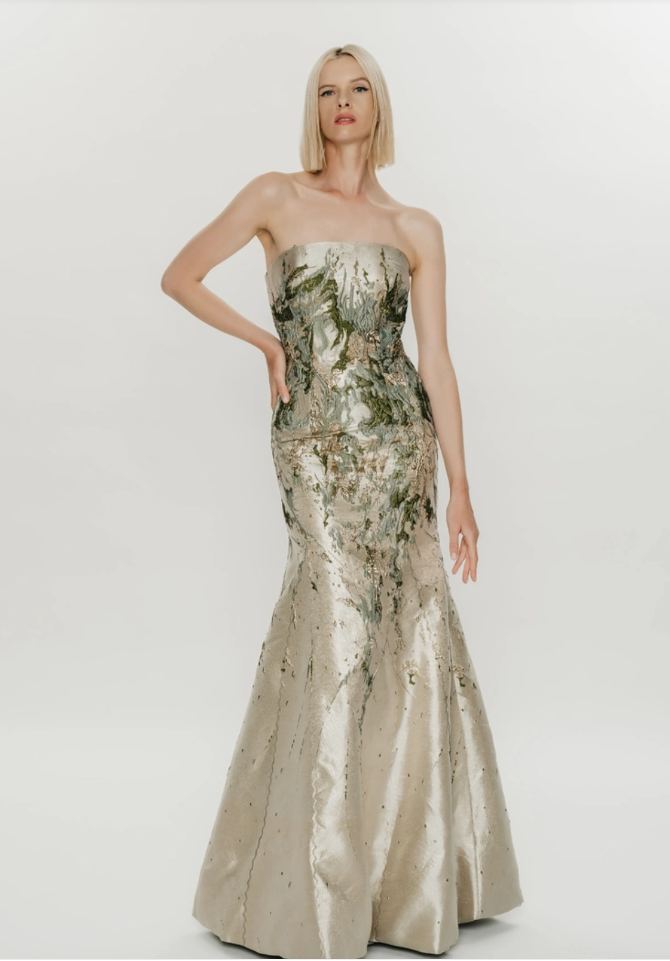 A model wearing a strapless, fitted gown with a shimmering gold and green design, featuring a mermaid silhouette. The dress has intricate detailing and sparkles, creating an elegant appearance.